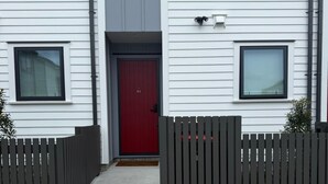 Una casa moderna de dos pisos con puerta roja, cerca de piquetes y un camino de concreto que lleva a la entrada.