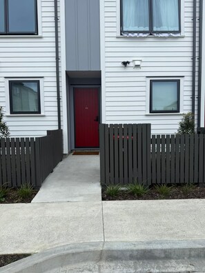 Une maison moderne à deux étages, avec une porte rouge, une clôture en bois et un chemin en béton menant à l’entrée.