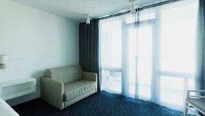 Habitación de hotel con sofá, una silla, una mesita de noche y un ventanal con cortinas.