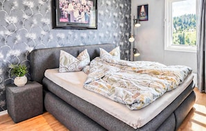 Ein Schlafzimmer mit grauem Schlafsofa, floralem Bettzeug, einem gerahmten Foto an der Wand, einer Stehlampe und einem Fenster mit Blick auf Bäume.