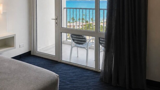 Ein Hotelzimmer mit Bett, Sessel, einem Balkon mit Meerblick und einem Vorhang vor dem Fenster.