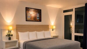 Une chambre d’hôtel avec un grand lit, des tables de chevet avec des lampes, un tableau au-dessus du lit et une porte-fenêtre coulissante sur le côté droit.