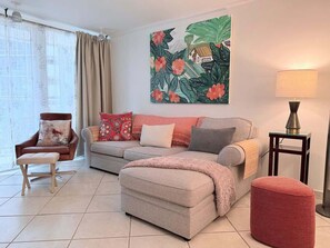 Ein Wohnzimmer mit Sofa, Hocker, Sessel, Lampe und einem Kunstwerk an der Wand.