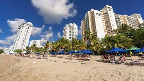 Ein Strand mit Palmen, Sonnenschirmen und Hochhäusern im Hintergrund.