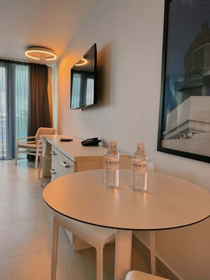 Ein Hotelzimmer mit einem runden Tisch, zwei Wasserflaschen, einem an der Wand montierten Fernseher, einem Sessel, einem Schreibtisch und einem Fenster mit Vorhängen.