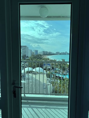 Blick von einem Balkon mit Geländer auf Strand, Palmen und eine Stadtlandschaft mit Gebäuden.