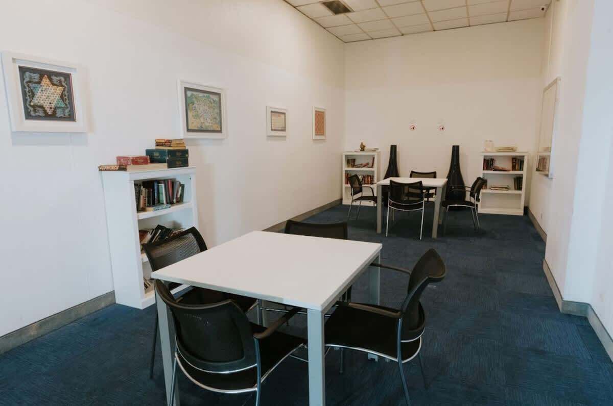 Een kamer met een witte tafel, zwarte stoelen, een boekenkast en lijstjes met afbeeldingen aan de muur.