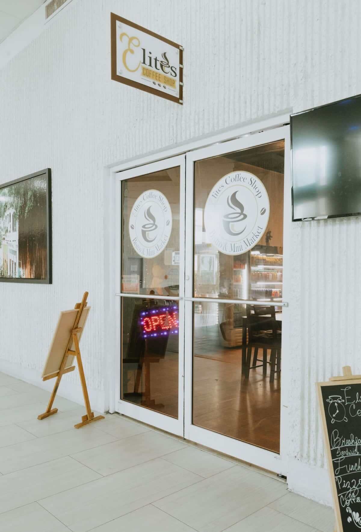 Una cafetería con un letrero que dice 'Elite's Coffee Shop', un letrero de neón que indica 'ABIERTO' y un menú en una pizarra en el exterior.
