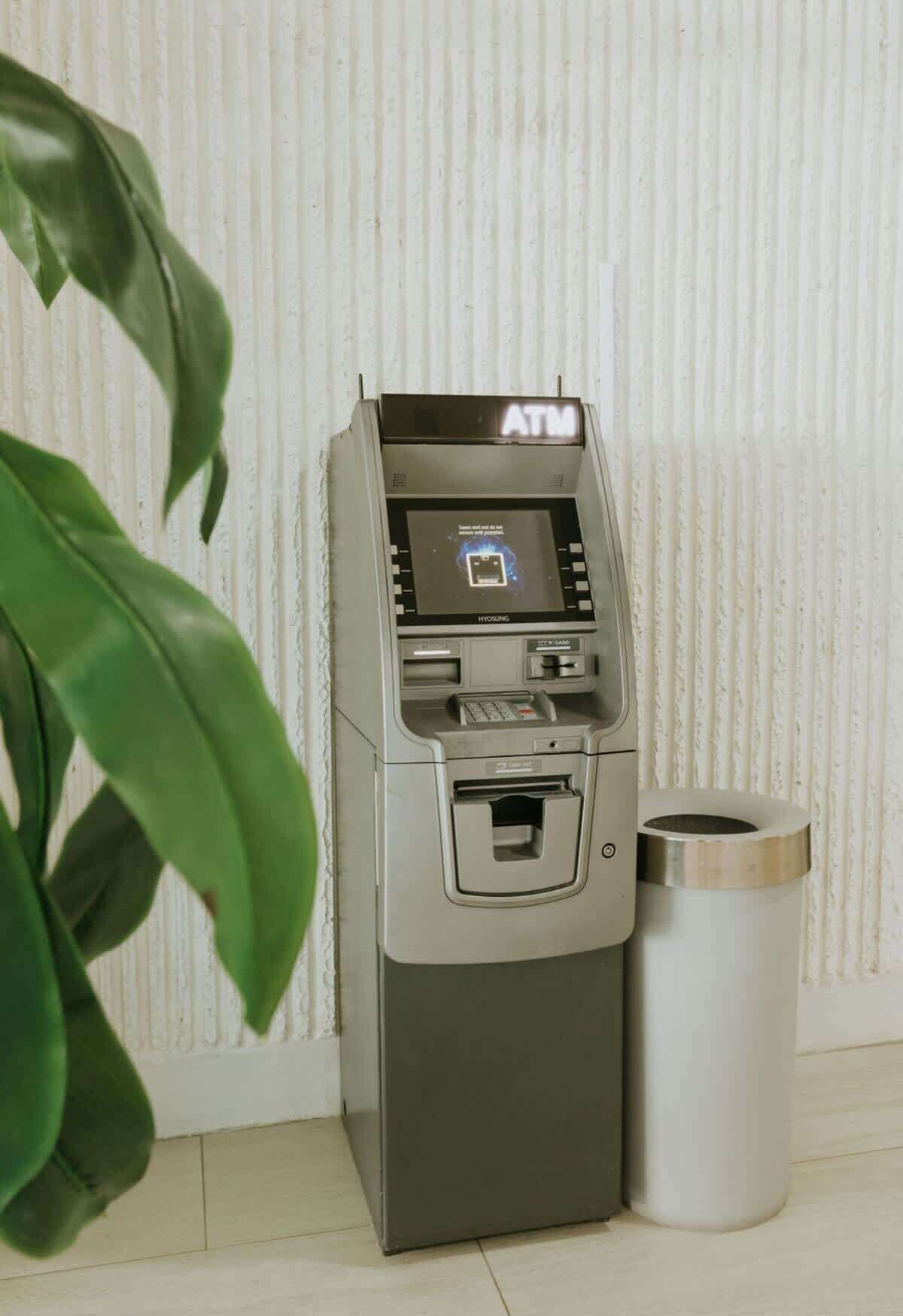Un distributeur automatique de billets, avec une poubelle à côté, adossé à un mur à texture.