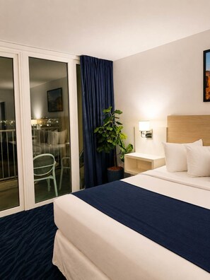 A hotel room with a bed, a nightstand, a lamp, a plant, and a view of the city through a glass door. - Ocean & Mountain View King Studio Isla Verde (Carolina)