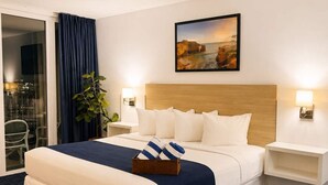 Una habitación de hotel con una cama grande, una mesita de noche, una lámpara de pared, una planta en maceta y un cuadro.