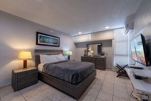 A modern hotel room with a bed, bedside tables, a desk, a TV, and a window with blinds. - 2BR w/King + 3 Twins • Balcony • on the Beach (Carolina)