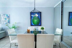 A modern dining area with a glass table, white chairs, a hanging light fixture, and artwork on the walls. - 2BR w/King + 3 Twins • Balcony • on the Beach (Carolina)