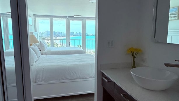 Habitación de hotel moderna con una cama grande, un baño con un espejo amplio y un lavamanos blanco, y un balcón con vistas a la ciudad.