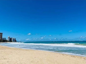 Ein Strand mit blauem Himmel, Meereswellen und Gebäuden im Hintergrund.
