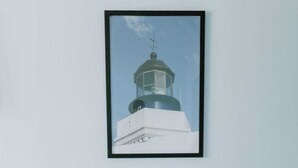 Un tableau encadré représentant un phare, accroché au mur, une table ronde blanche avec deux chaises et un mur de couleur bleu clair.
