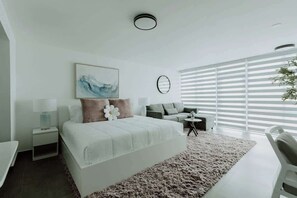 Ein modernes Schlafzimmer mit einem großen Bett, einer Couch, einem kleinen Tisch und einem großen Fenster mit Jalousien.