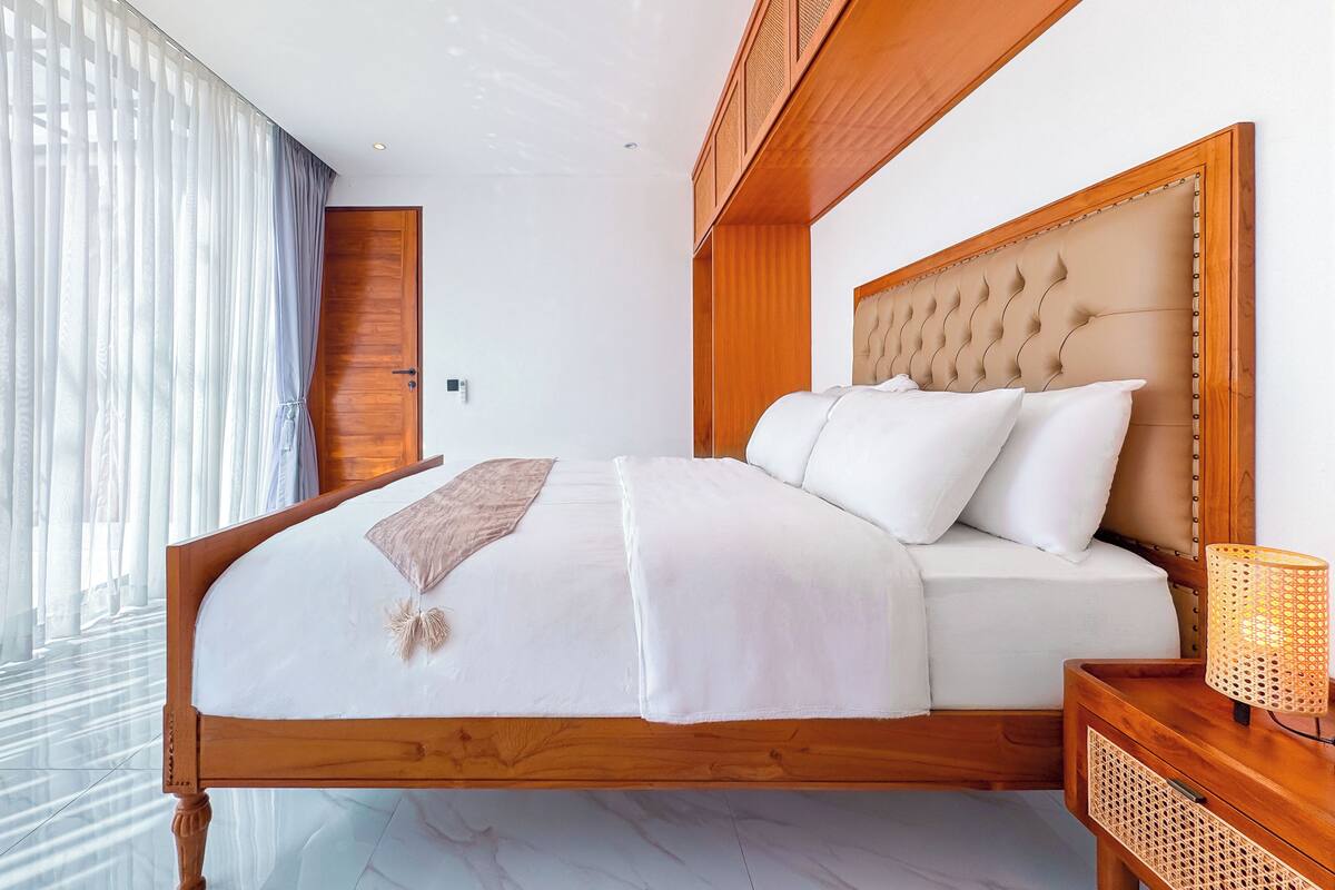 A bedroom with a wooden bed frame, a headboard, a bedside table with a lamp, and a window with sheer curtains.