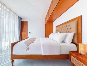 A bedroom with a wooden bed frame, a headboard, a bedside table with a lamp, and a window with sheer curtains. - Villa Exquisite Seminyak by Nagisa Bali  (Badung)
