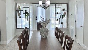 Una sala da pranzo con un lungo tavolo in legno, sedie, un lampadario e piante in vasi su mensole.
