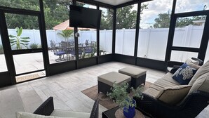 Una veranda coperta con mobili in vimini, una televisione a schermo piatto e vista su un giardino recintato da una staccionata bianca con mobili da esterno.