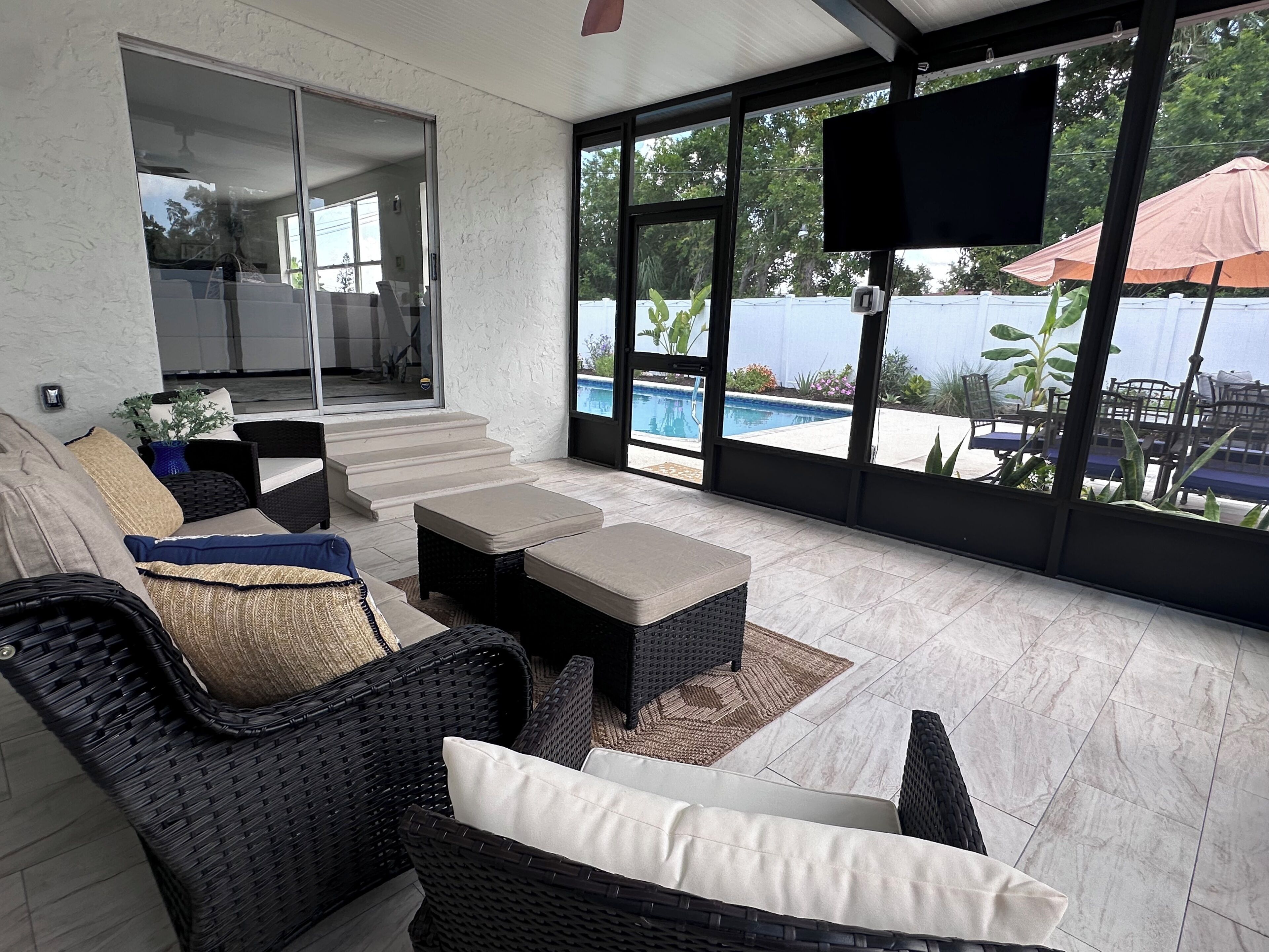 A covered outdoor seating area with wicker furniture, a flat-screen TV, and a view of a swimming pool.