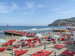 Ein Strand mit roten Liegestühlen und gestreiften Sonnenschirmen, ein Steg mit roten Stühlen sowie der Blick auf das Meer und Gebäude im Hintergrund.