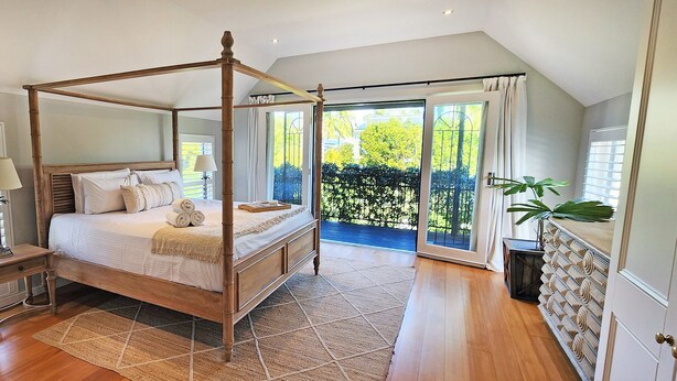 A four-poster bed with a wooden frame, a bedside table with a lamp, a large window with blinds, and a view of greenery outside.