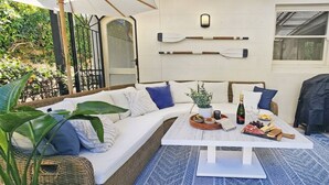 A cozy outdoor seating area with a white sofa, blue cushions, a wooden table with a bottle, glasses, and snacks, and a covered grill.