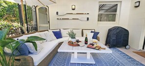 Eine gemütliche Sitzgruppe im Freien mit einem weißen Sofa, blauen Kissen, einem Holztisch mit einer Flasche, Gläsern und Snacks sowie einem überdachten Grill.