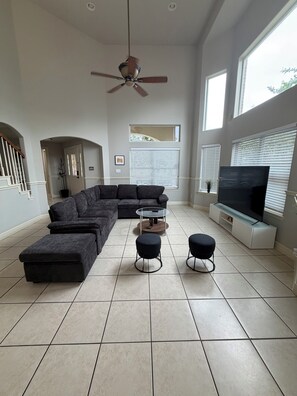 A modern living room with a sectional sofa, ottomans, a glass coffee table, a ceiling fan, a flat-screen TV, and large windows.