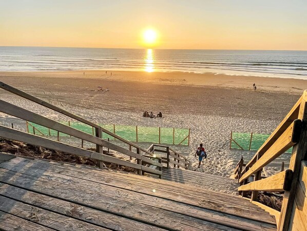 Eine Holztreppe führt zu einem Strand, im Hintergrund ist ein Sonnenuntergang zu sehen.