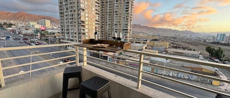 A balcony with a table, two stools, and a view of a cityscape with high-rise buildings and mountains in the background.