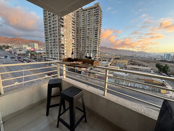 Ein Balkon mit Tisch, zwei Hockern und Blick auf eine Stadtlandschaft mit Hochhäusern und Bergen im Hintergrund.