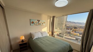 A hotel room with a bed, a nightstand, a lamp, a painting, and a large window with a city view.