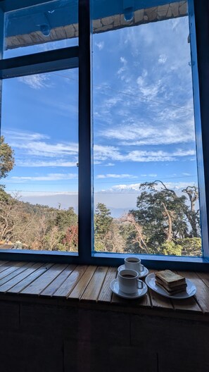Ein Fenster mit Blick auf eine gebirgige Landschaft, ein Holztisch mit einer Kaffeetasse und einer Scheibe Toast.