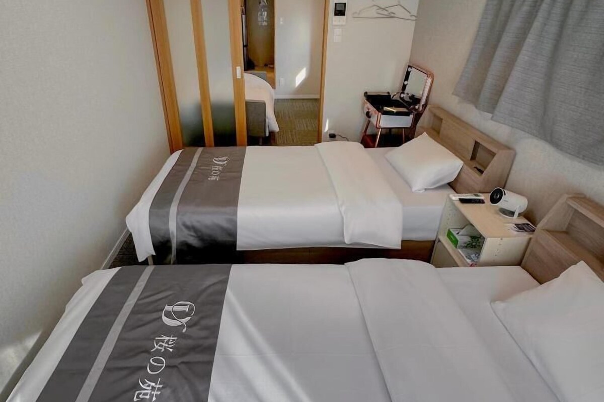 Een hotelkamer met twee bedden, een nachtkastje met lamp, een stoel en uitzicht op de gang.