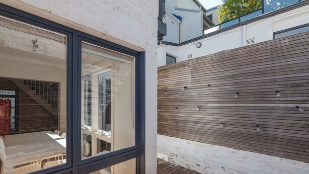 Een modern buiten terras met een houten dek, een glazen omheining en een witte bakstenen muur.