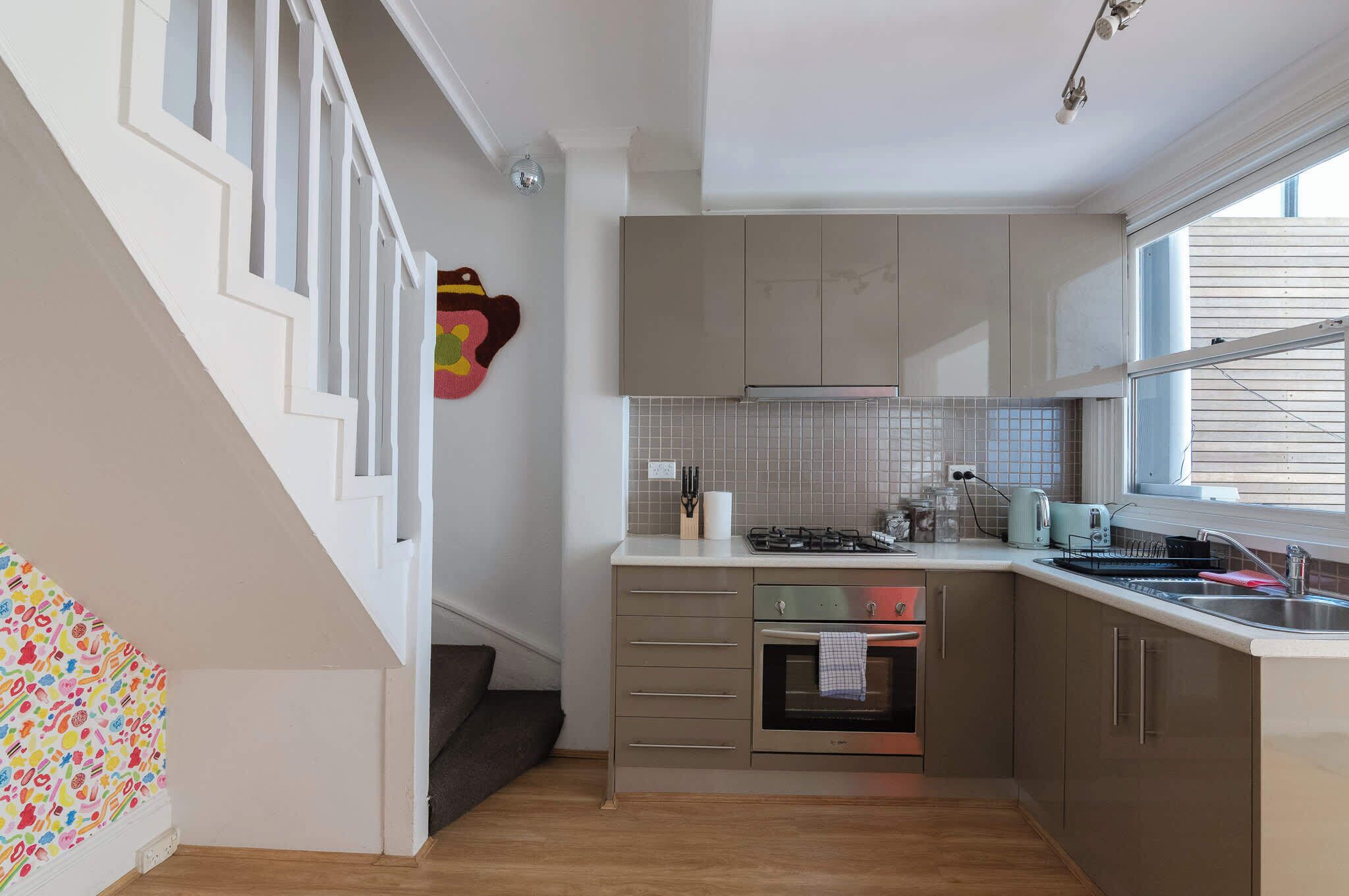 A kitchen with a stove, oven, and sink, a staircase with white railings, and a window with blinds.