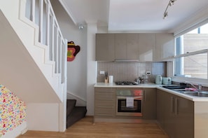 Eine Küche mit Herd, Backofen und Spüle, eine Treppe mit weißen Geländern sowie ein Fenster mit Jalousien.