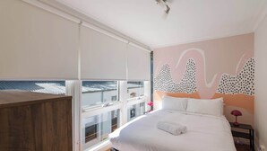 Een slaapkamer met een groot bed, een houten hoofdbord, een raam met zonwering, een muur met abstracte kunst en een nachtkastje met een lamp.