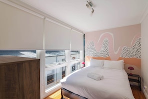 Ein Schlafzimmer mit einem großen Bett, einem Holzkopfende, einem Fenster mit Jalousien, einer Wand mit abstrakter Kunst und einem Nachttisch mit einer Lampe.