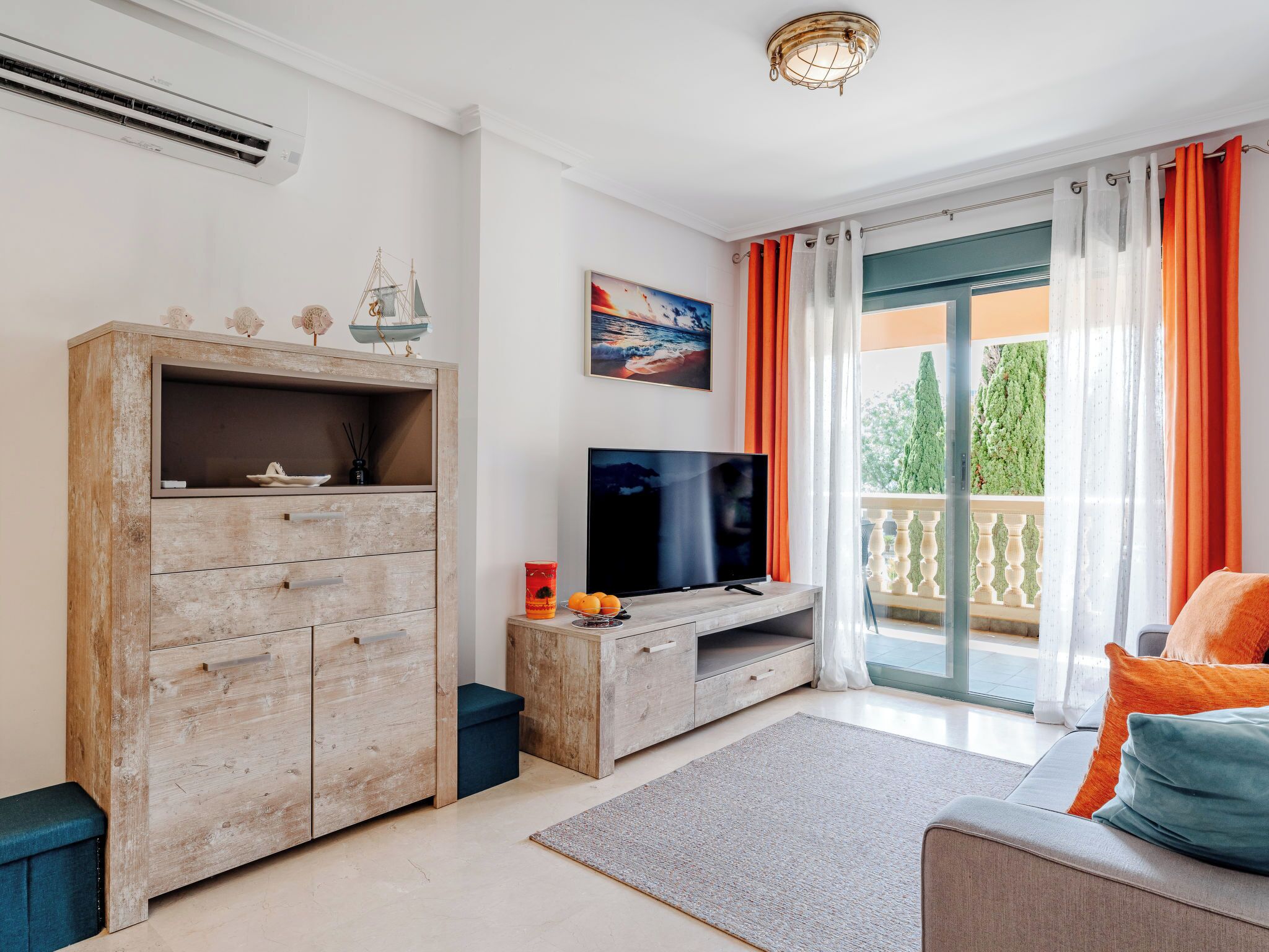 Una sala de estar con un mueble de madera, un televisor de pantalla plana, un sofá con cojines naranja y un balcón con cortinas anaranjadas.