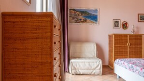 A bedroom with a wooden dresser, a white chair, a bed with a floral bedspread, and a painting of a coastal scene. - Bilocale la Marea - Goelba (Portoferraio)