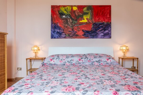Et soveværelse med blomstret sengetæppe, to sengeborde med lamper, og et stort abstrakt billede på væggen.