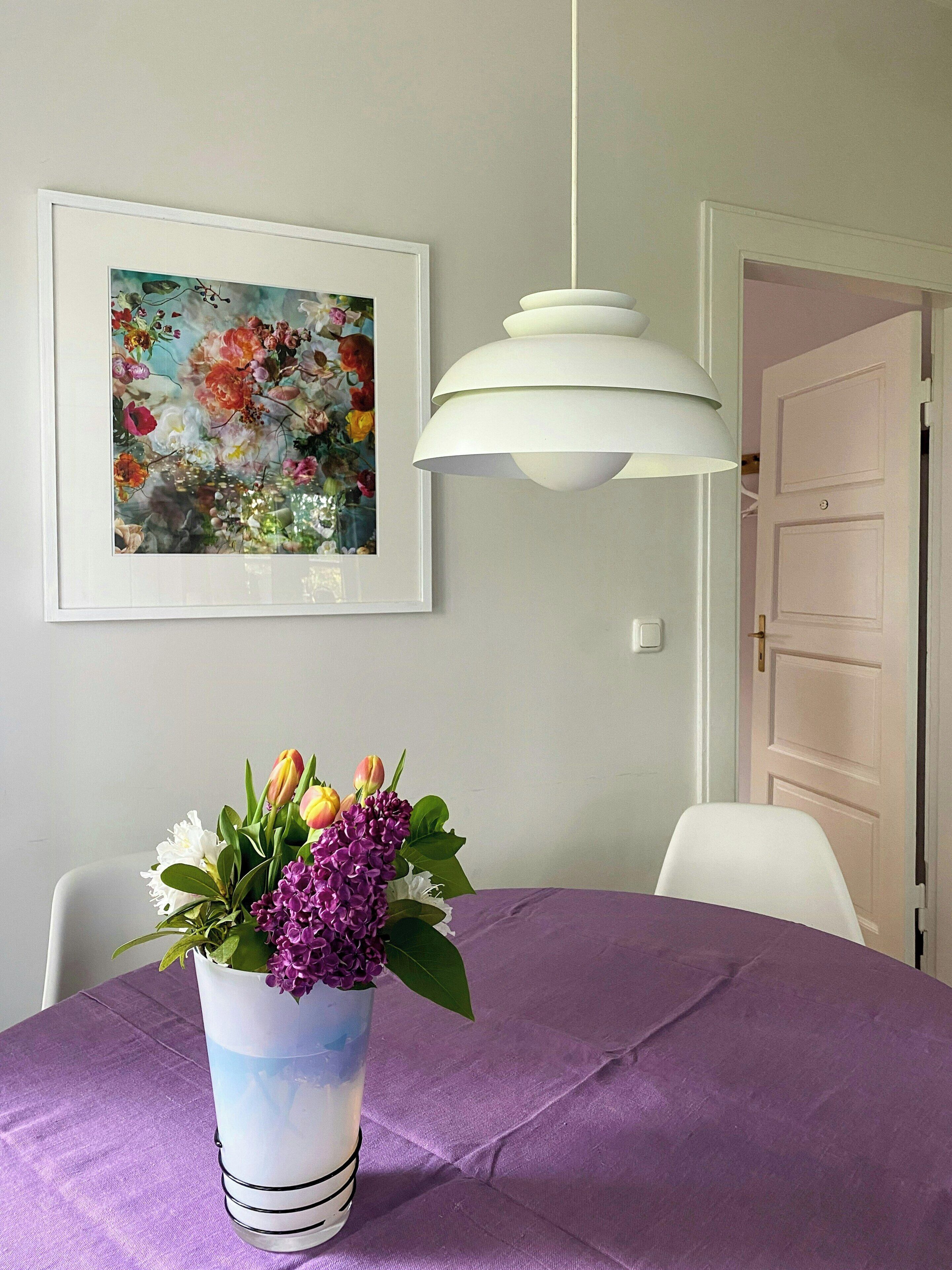 Un coin repas avec une nappe violette, un vase rempli de fleurs, un luminaire suspendu blanc et un tableau encadré accroché au mur.