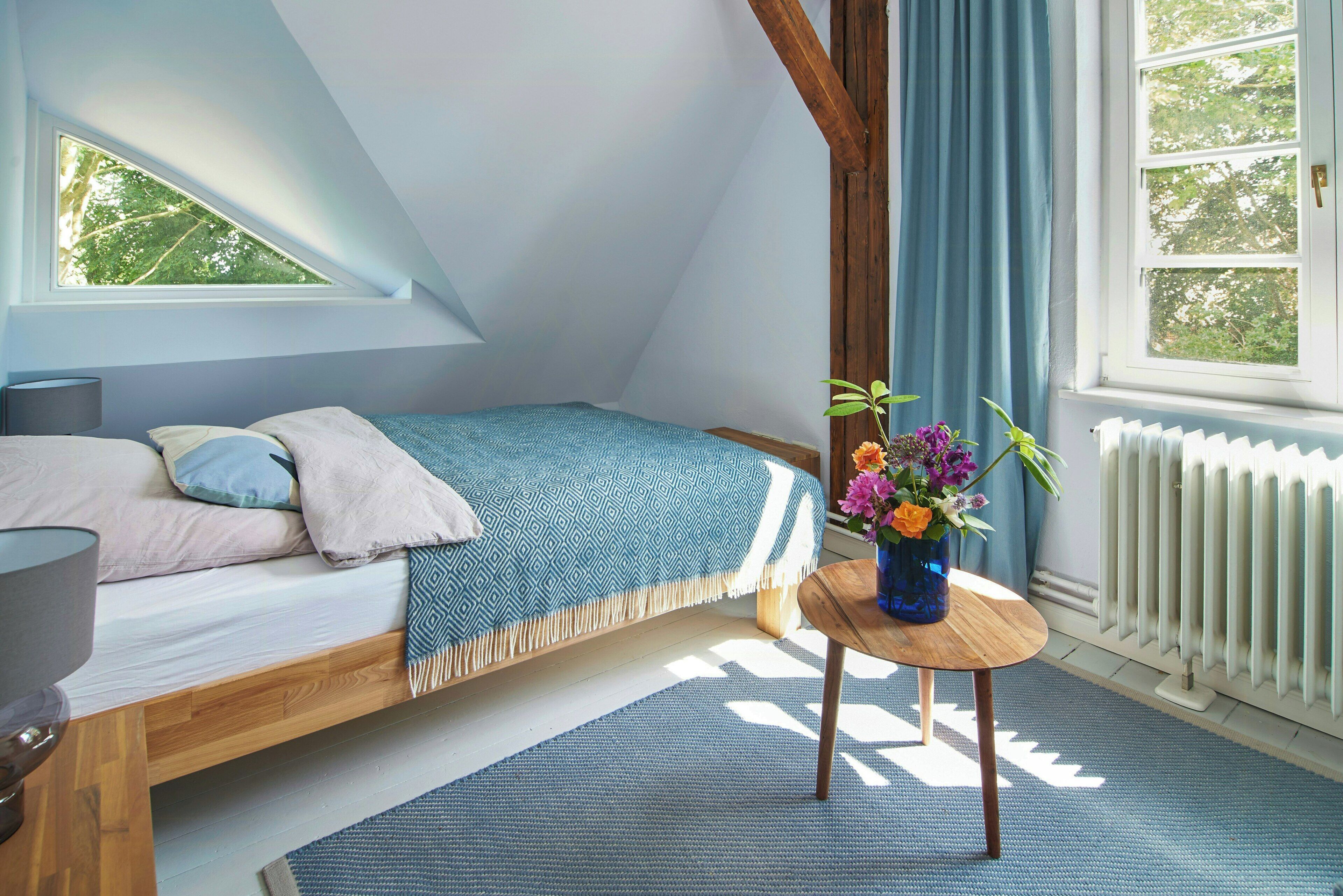 Ein Schlafzimmer mit schrägen Wänden, einem Bett mit gemustertem Überwurf, einem Nachttisch mit Blumenvase und einem Fenster mit Vorhängen.