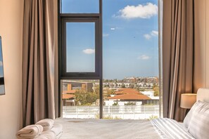 A hotel room with a large window, a bed with white bedding, and a view of the cityscape. - Bright 2BR Apartment in Central Bang Tao (Choeng Thale)