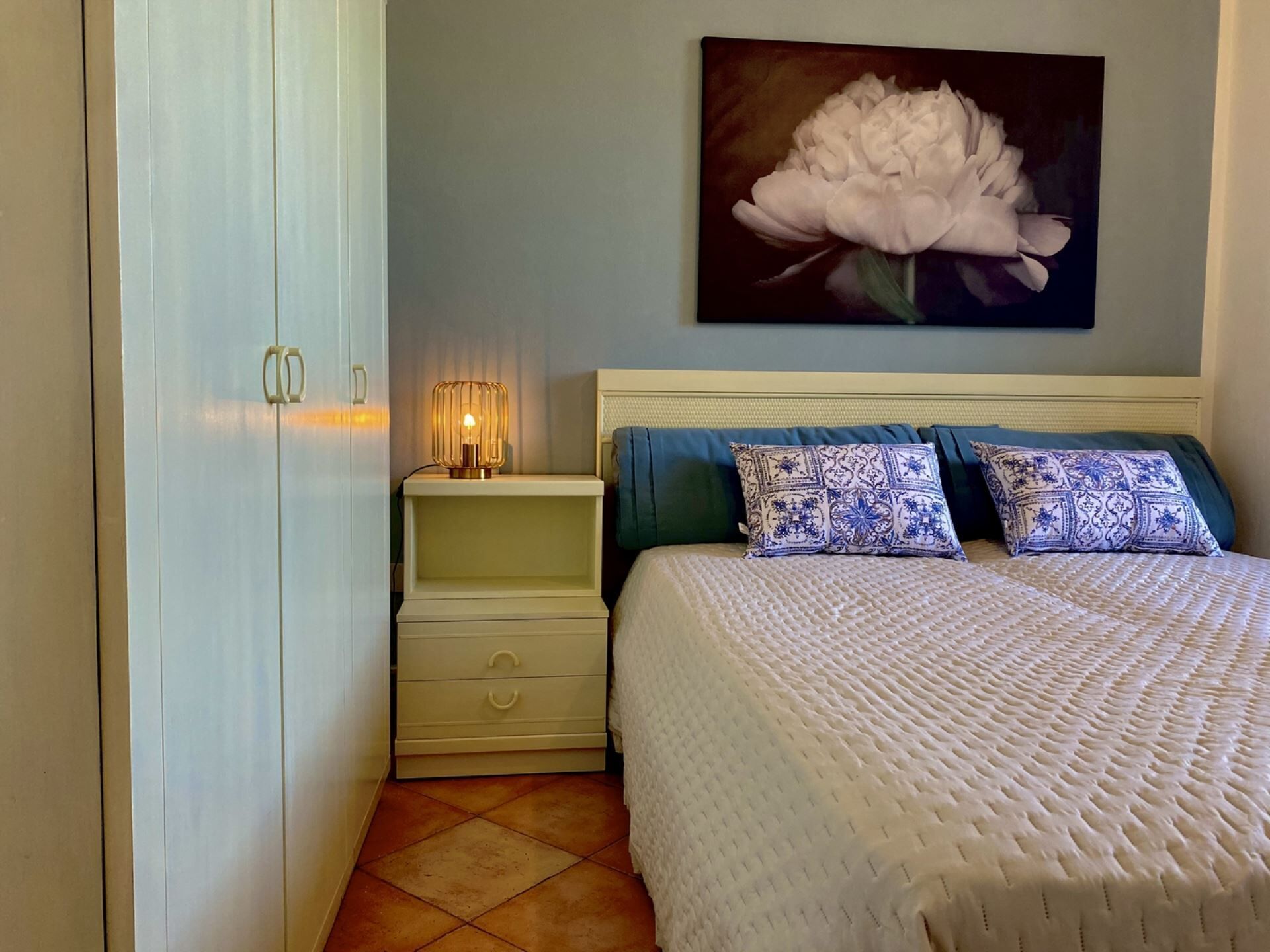 Een slaapkamer met een bed, nachtkastje, lamp en een schilderij aan de muur.