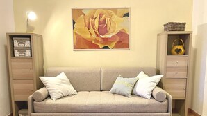 Um sofá em tons neutros, com três almofadas, ladeado por armários de madeira com gavetas e prateleiras, em frente a uma parede lisa com um quadro abstrato emoldurado.